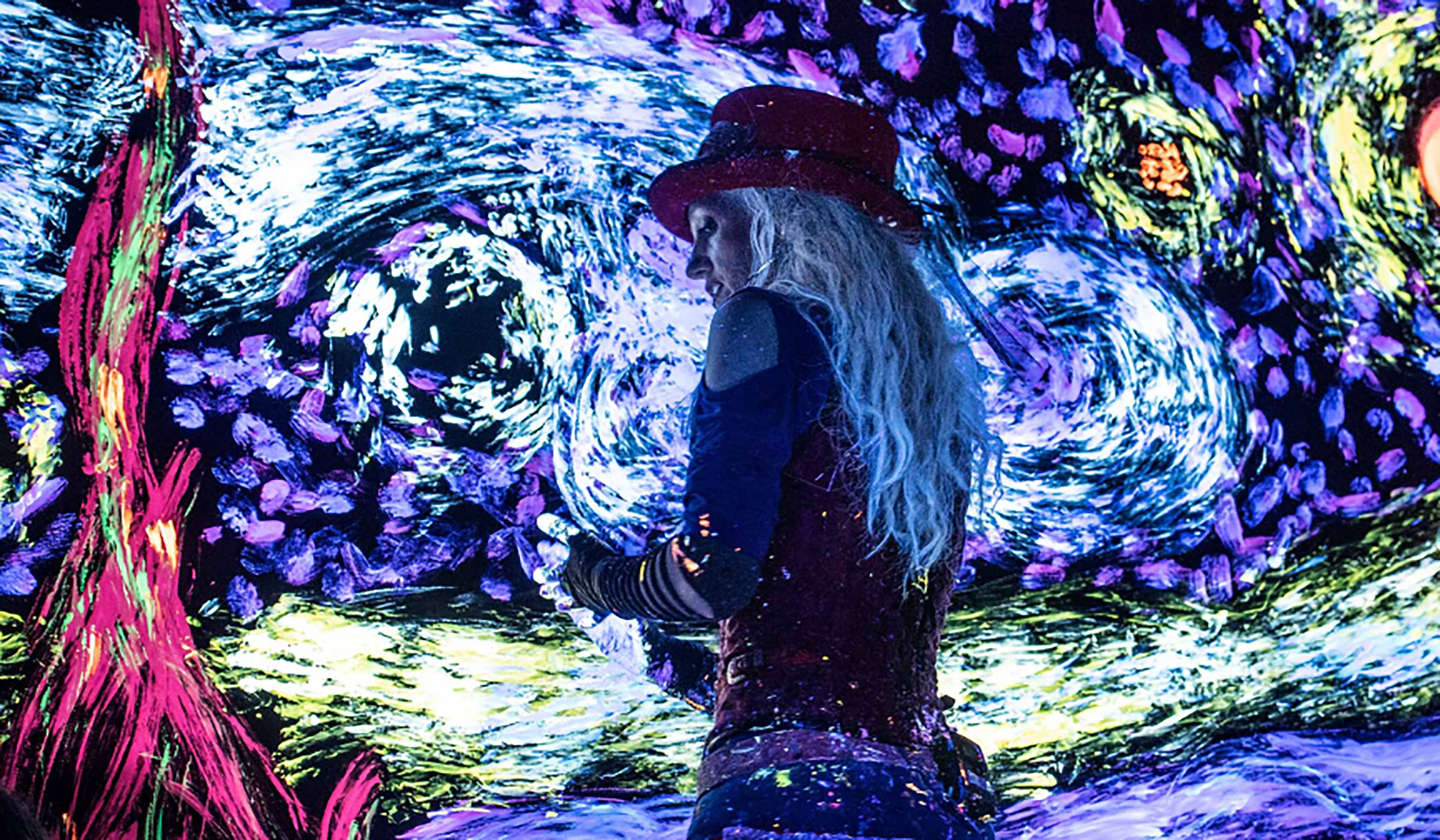 A woman wearing a top-hat paints on a black canvas in a dark room using black light paint that glows in the dark.